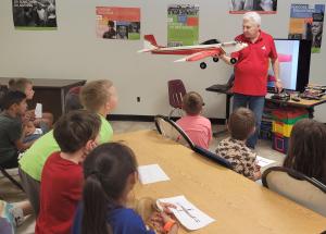 Educator shows model airplane to attentive children in classroom.