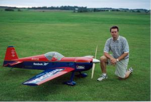 Man kneeling beside a large remote-controlled airplane on a grassy field.