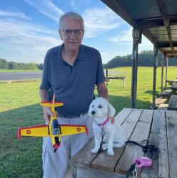Elderly man smiling, holding a model plane, with a small white dog on a picnic table.