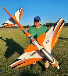 Man holding large orange and white model jet on grassy field.