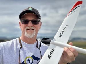 Man with cap smiling, holding a model glider airplane outdoors.