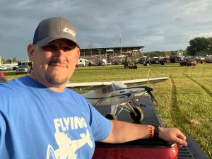 Man in blue shirt with a model airplane on a truck, outdoor event in background.