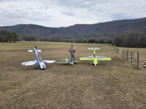 Man standing with three model aircraft on a field, mountains in the background.