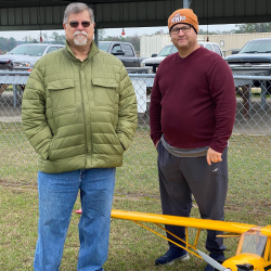 Two men standing by a yellow model airplane, under a sheltered area.
