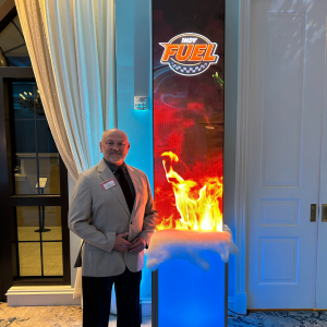 Man in gray suit stands beside digital display with Indy Fuel logo and flame graphics.