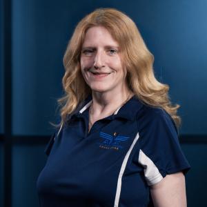 Smiling woman with wavy red hair in a navy polo shirt against a dark blue background.