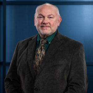 A man in a brown suit and tie, standing against a dark blue background.