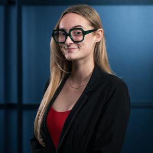 Woman with long hair, wearing glasses and a black blazer, against a blue background.