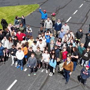 Large group of people gathered outdoors on a paved area, surrounded by green grass.