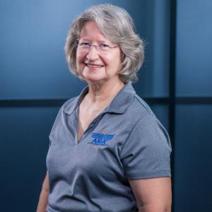 Smiling woman in a gray polo shirt stands against a blue background.
