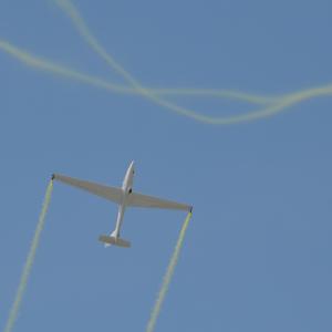White glider soaring with yellow smoke trails against blue sky.