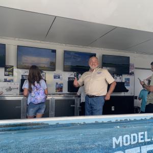 Man in beige shirt giving thumbs up, standing near display monitors indoors.