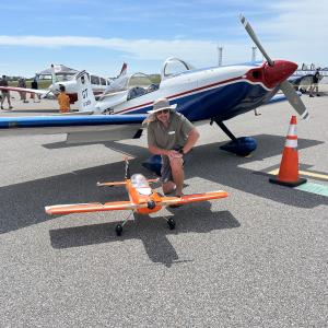 Man kneeling beside an orange model plane, with a larger blue and white plane behind.