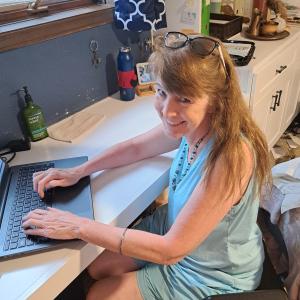 Smiling woman in blue dress typing on a laptop in a home office.