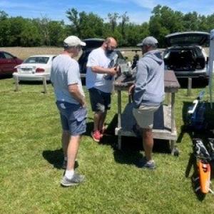 Three men standing by a table with equipment outdoors on a sunny day.