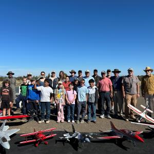 A group of people posing with model airplanes outside on a sunny day.