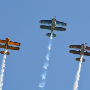 Three biplanes with smoke trails perform aerobatics against a clear blue sky.