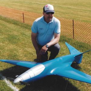 Man kneeling beside a large blue model jet on a grassy field.