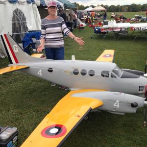 A woman stands beside a large model airplane with yellow wings on a grassy field.