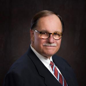 Man in a suit with glasses, striped tie, and mustache against dark background.