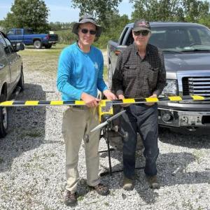 Two people holding a model plane, standing between parked trucks on gravel.