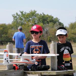 Two boys with a model airplane outdoors, wearing hats and sunglasses.
