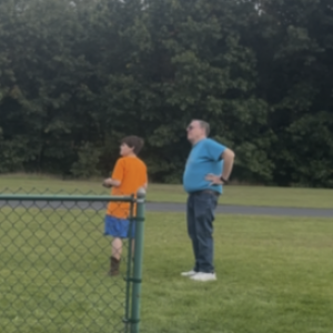 Two people standing on grass near a fence, trees in the background.