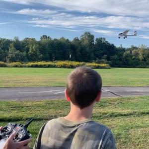 Boy watching remote-controlled plane flying in a grassy field.
