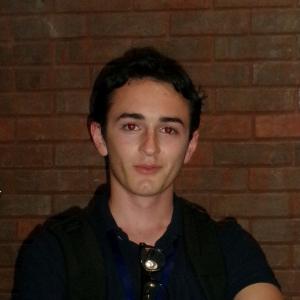 Young man with dark hair stands against a brick wall background.