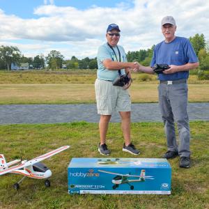 Two men shaking hands near a model airplane on grass with a plane box.