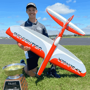 Young person kneeling on grass, smiling with a model airplane and a trophy.