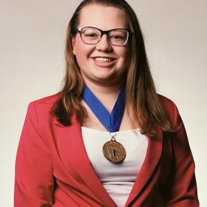 Smiling person in a red blazer and glasses, wearing a gold medal on a blue ribbon.