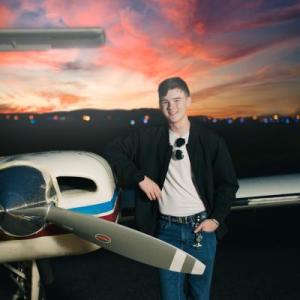 Young person smiling, leaning on small airplane at sunset.