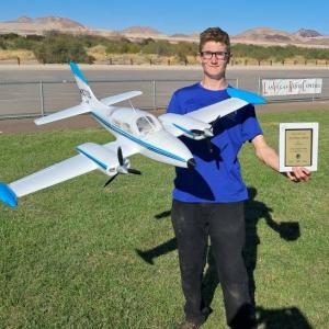 Man holding large model airplane and award, standing on grass near runway.
