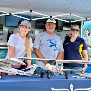 Three people smiling at a booth with model airplanes on display.
