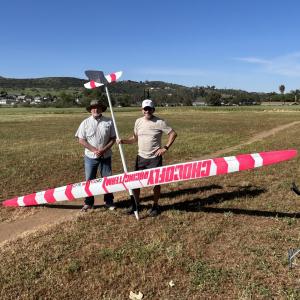 Two people smile next to a large pink and white model glider in an open field.