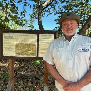 Man in a hat stands smiling by an informational sign in a wooded area.