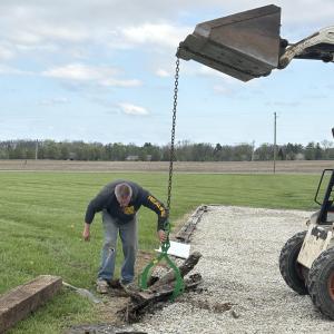 A man guiding a metal pipe being moved by an excavator on a gravel path.