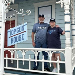 Couple on a porch with a sign: "Top Gun House."
