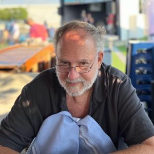 Man with glasses and a beard sitting outside at a table, wearing a dark shirt.