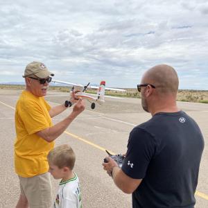 Two men and a child with model airplane on airstrip, cloudy sky.