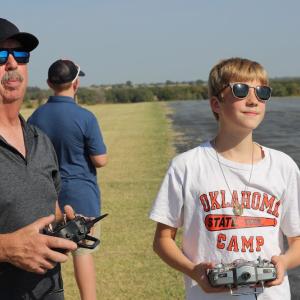 Two people controlling model aircraft outdoors, one wearing sunglasses and a cap.