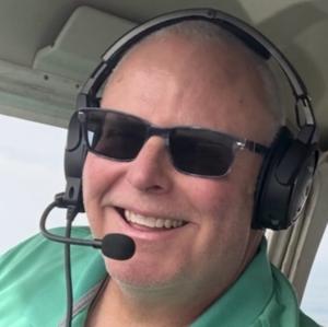 Man smiling in cockpit, wearing sunglasses, headset, and green shirt.