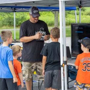 Man showing remote control to four kids near a monitor under a tent.