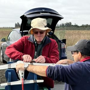 Two people working on a model airplane outdoors, near a parked car.