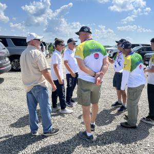 Group of people standing on a gravel lot under a blue sky, talking and wearing casual clothes.