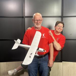 Man and woman in red shirts smiling, holding a white model airplane.