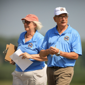 A man and woman wearing blue polo shirts and caps stand outdoors, holding a clipboard and papers.