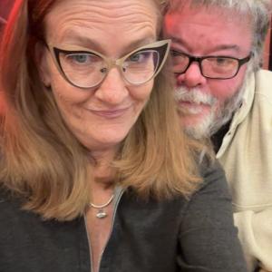 Two people wearing glasses, smiling in a warmly lit setting.