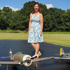 Woman standing on model airplane outdoors, wearing a floral dress, trees in the background.